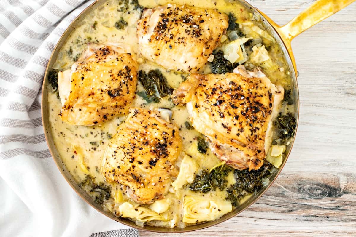 Baked chicken thighs in a skillet with kale and artichoke sauce.