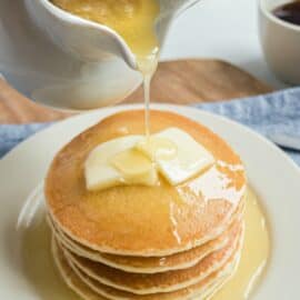 Buttermilk syrup in a white pitcher being poured over a stack of pancakes.