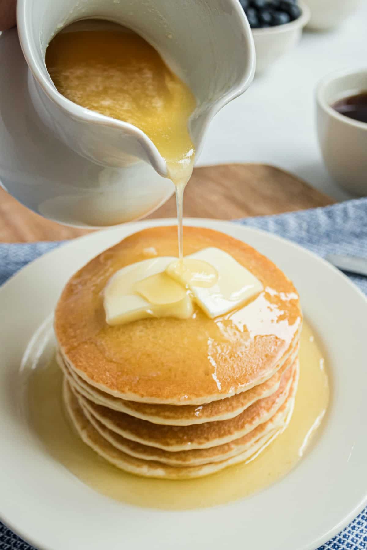 Buttermilk syrup in a white pitcher being poured over a stack of pancakes.