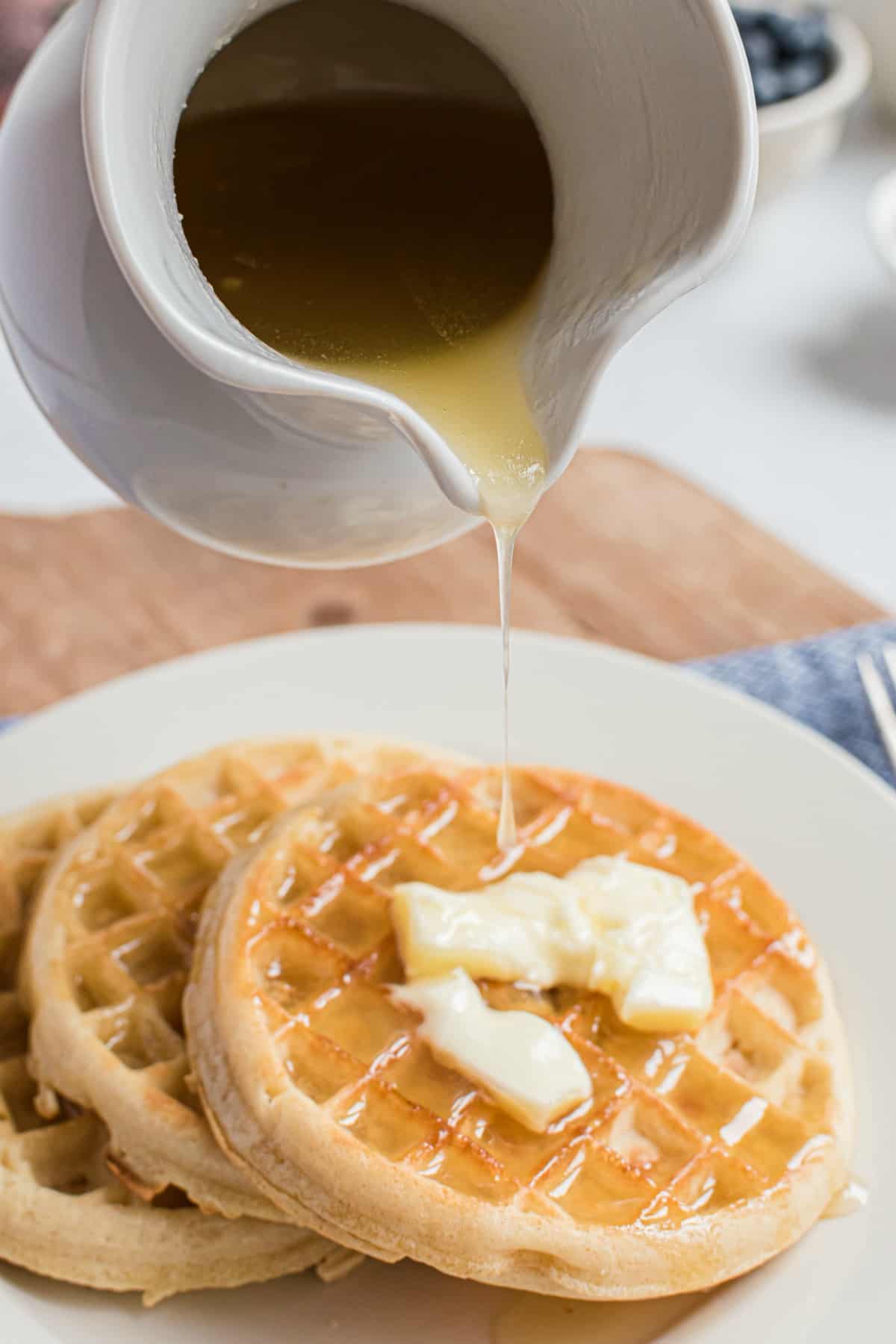 Homemade buttermilk syrup in a white pitcher being poured over a stack of homemade waffles.