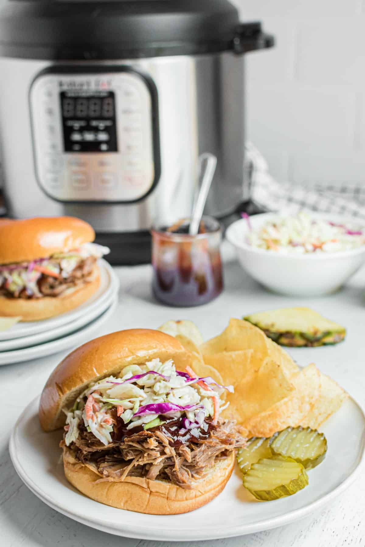 Hawaiian pork on a bun with bbq sauce and coleslaw with a side of chips and pickles. Instant pot in background.