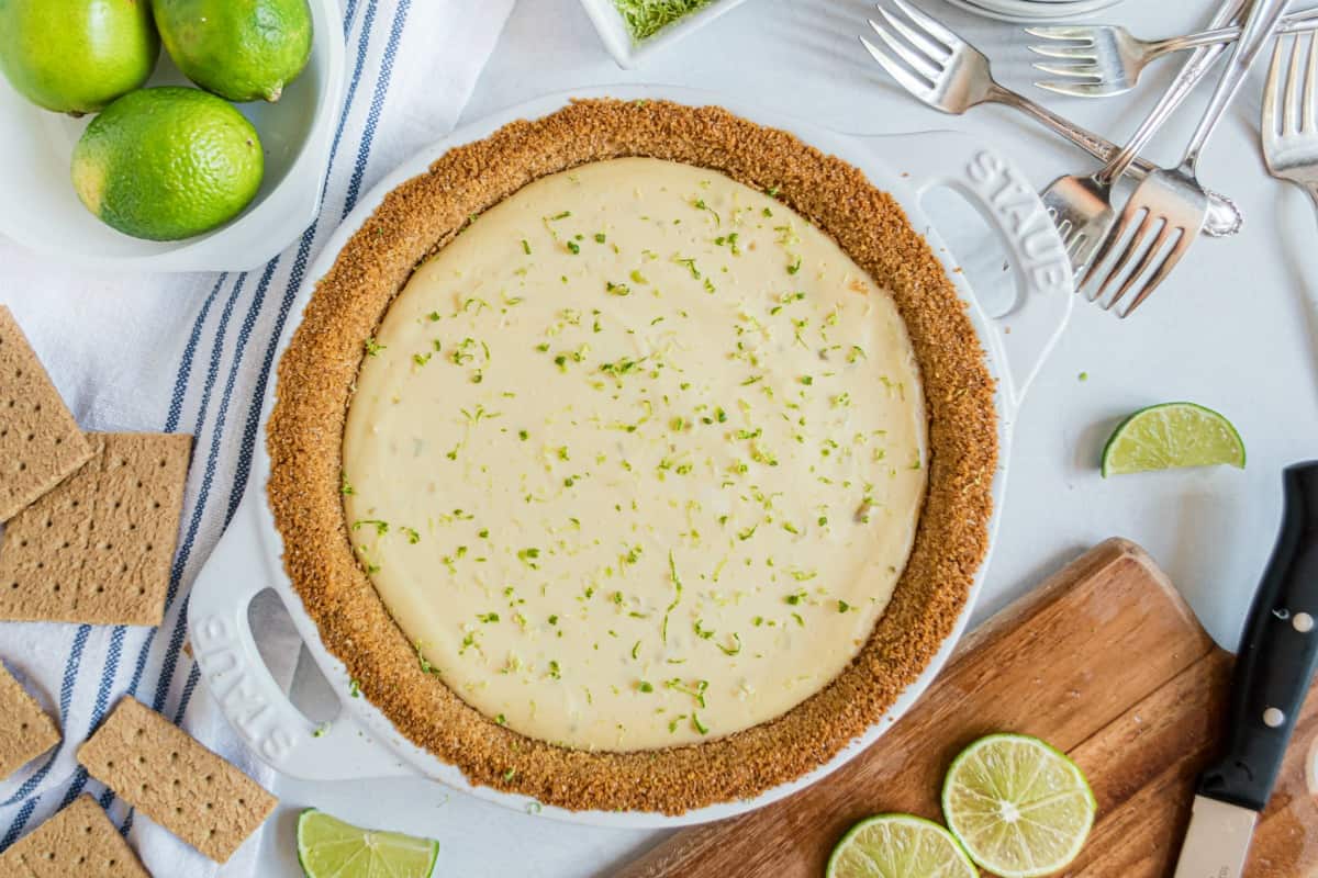 Key lime pie in a white pie plate, with limes, graham crackers, and a cutting board in background.