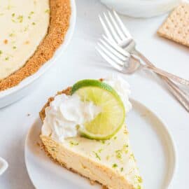 Slice of key lime pie on a white plate and garnished with whipped cream, lime zest, and a slice of lime.