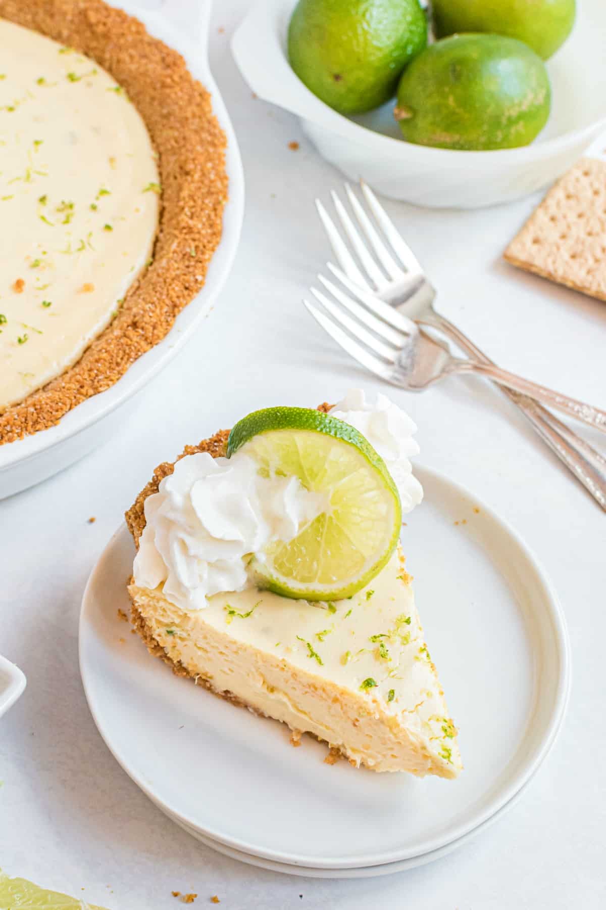 Slice of key lime pie on a white plate and garnished with whipped cream, lime zest, and a slice of lime.