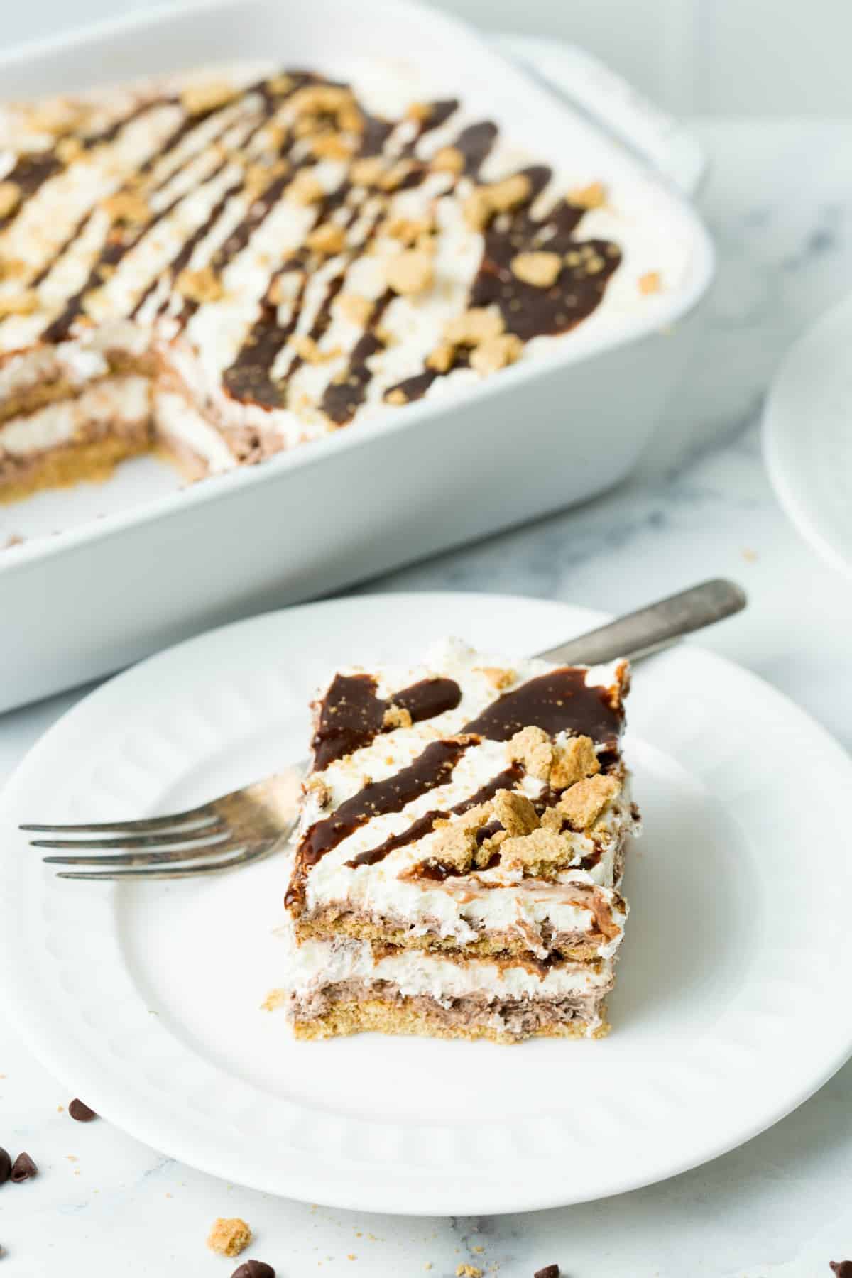 Slice of smores cake on a white plate with silver fork. Dish of cake in the background.