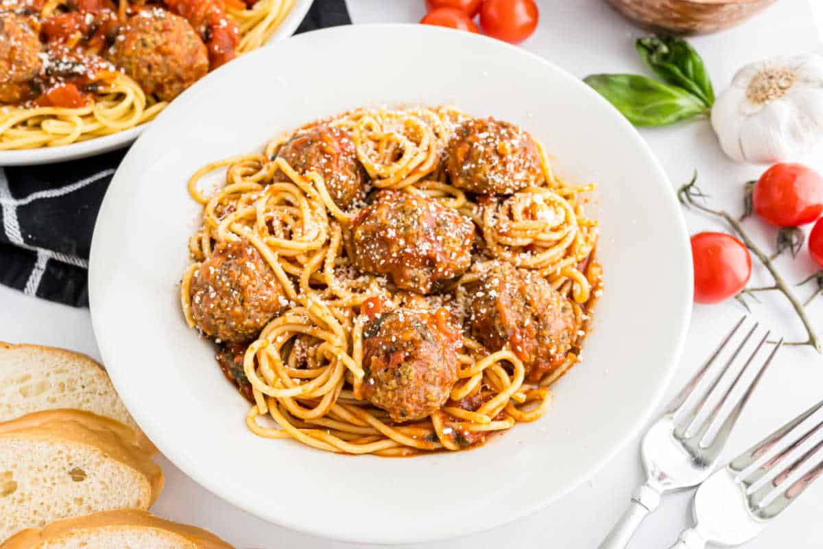 A bowl of spaghetti with turkey meatballs in bruschetta sauce, topped with grated cheese, surrounded by bread and fresh ingredients.