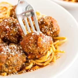 A fork holds a turkey meatball with bruschetta sauce over spaghetti topped with tomato sauce and grated cheese on a white plate.