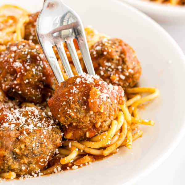 A fork holds a turkey meatball with bruschetta sauce over spaghetti topped with tomato sauce and grated cheese on a white plate.