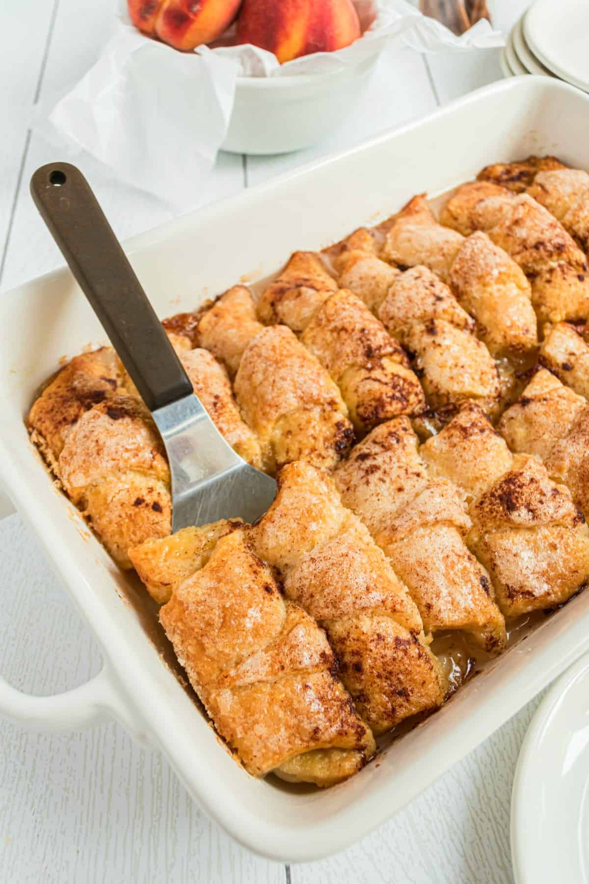 Peach dumplings baked in a 13x9 white baking dish and being removed with spatula.