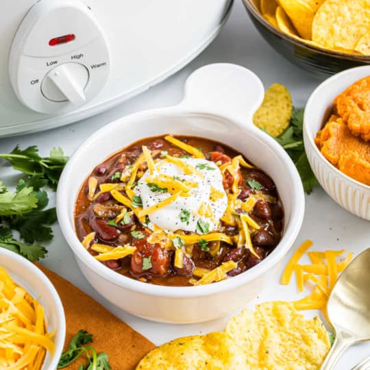 Bowl of pumpkin chili topped with sour cream and cheese, surrounded by chips, cheese, cilantro, and a slow cooker.