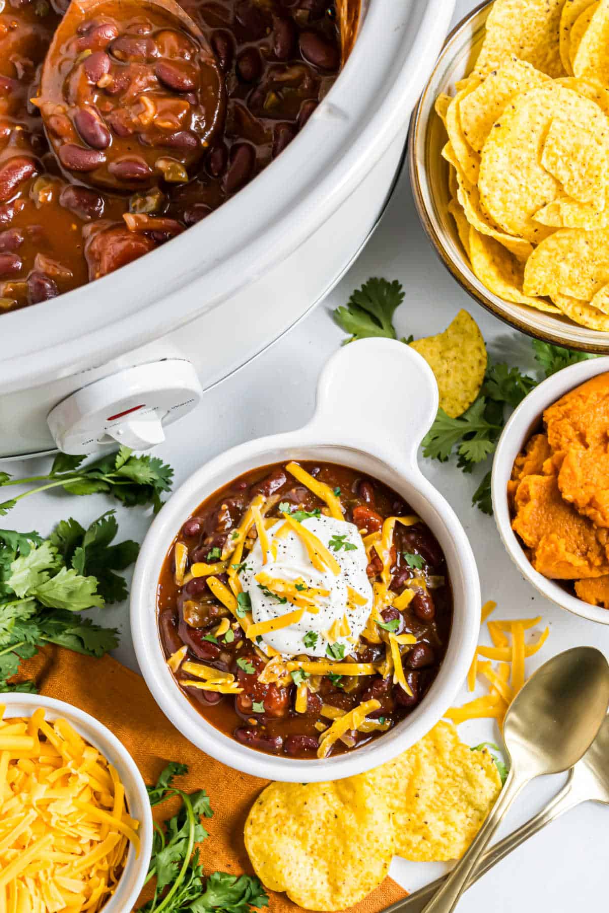 Bowl of pumpkin chili topped with sour cream and cheese, surrounded by tortilla chips and garnishes.