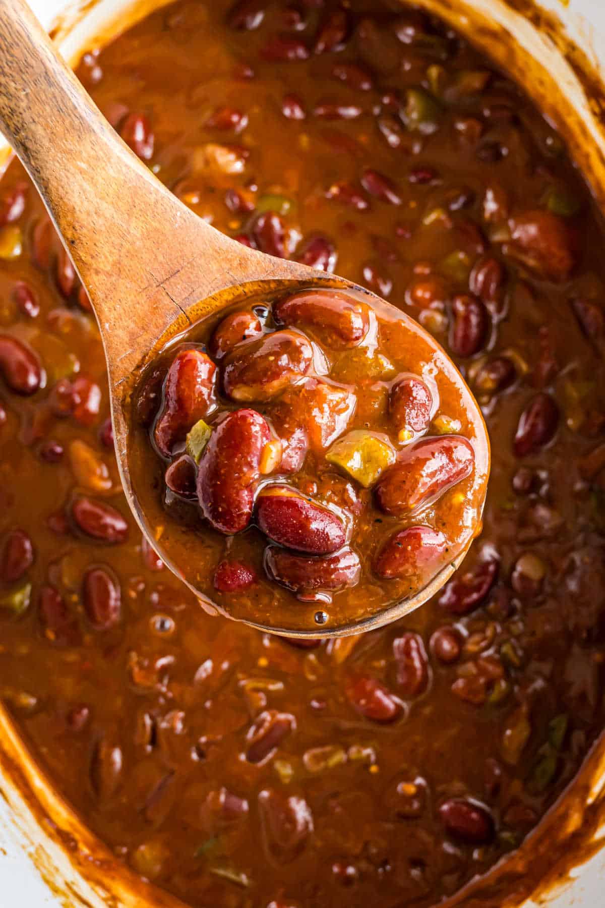 A wooden spoon scoops up hearty pumpkin chili with red kidney beans and vegetables from a pot.