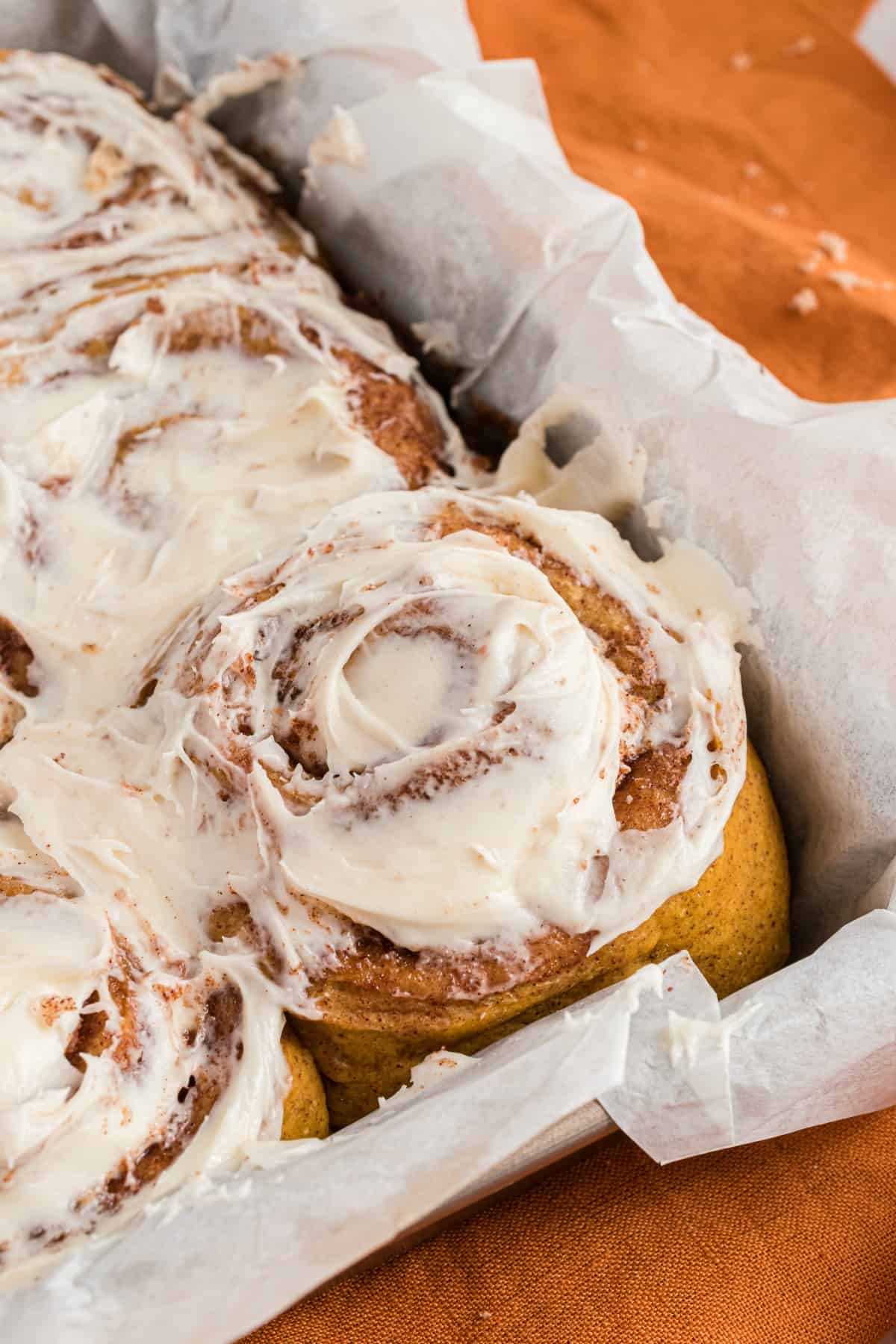 Pumpkin cinnamon rolls in parchment paper lined baking dish with maple icing.