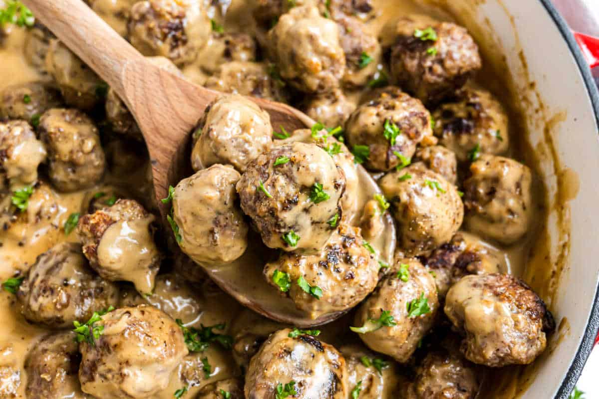 Swedish meatballs covered in creamy sauce, garnished with parsley, in a pot with a wooden spoon scooping some.