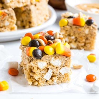 A peanut butter candy corn krispie treat topped with candy corn and chocolate candies sits on parchment paper.