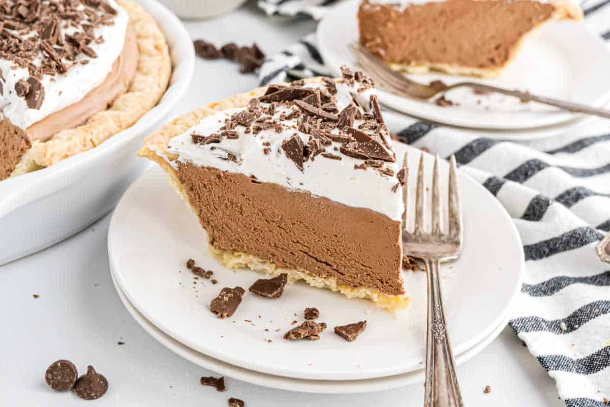 A slice of decadent french silk pie topped with whipped cream and chocolate shavings sits on a white plate with a fork.