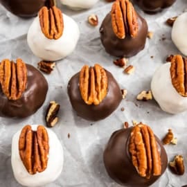Chocolate and white-coated pecan pie truffles topped with pecan halves rest on parchment paper, surrounded by scattered pecan pieces.
