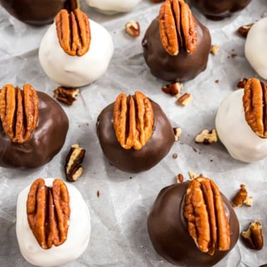 Chocolate and white-coated pecan pie truffles topped with pecan halves rest on parchment paper, surrounded by scattered pecan pieces.