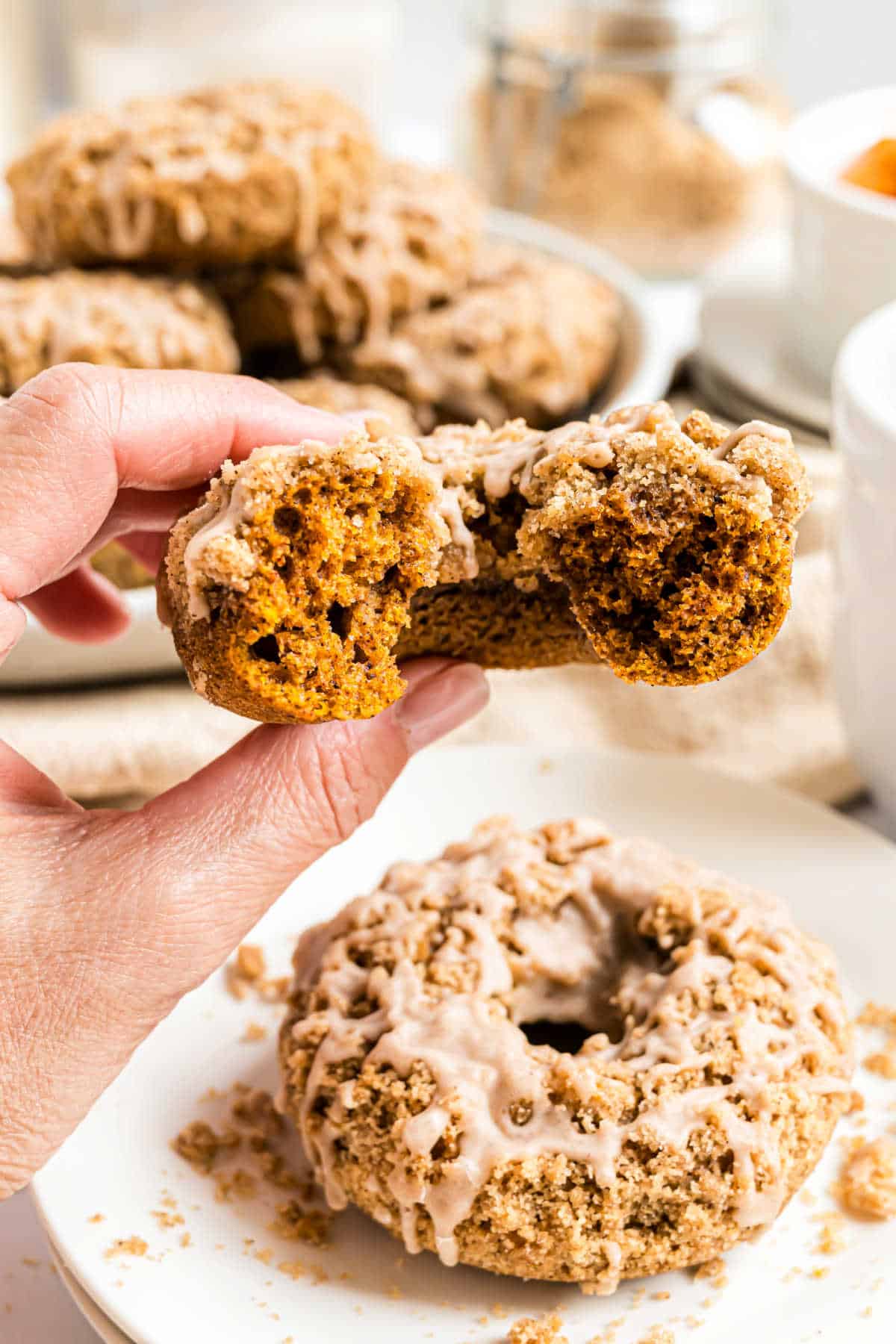 A hand holds a pumpkin streusel donut with cinnamon glaze, revealing the moist inside; more donuts rest on a plate below.