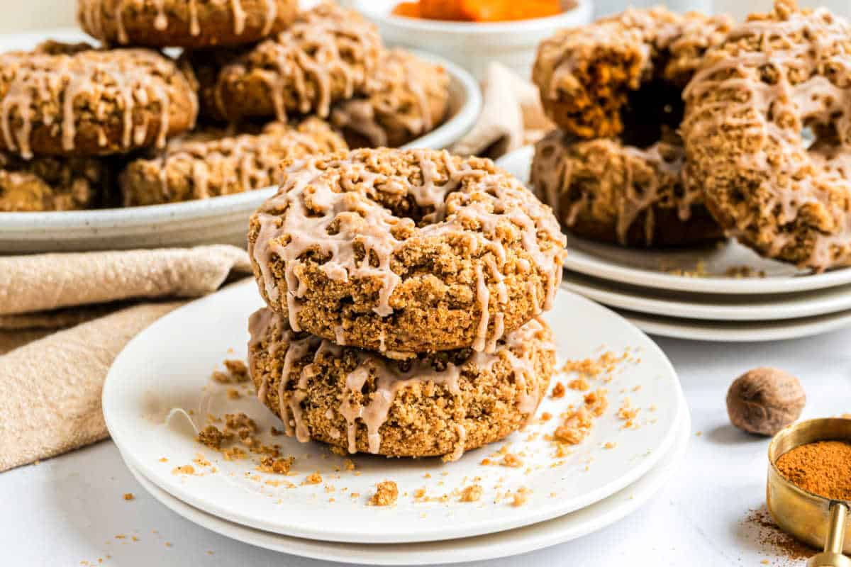 Two crumb-topped pumpkin streusel donuts with icing are stacked on a plate, with more donuts and ingredients in the background.