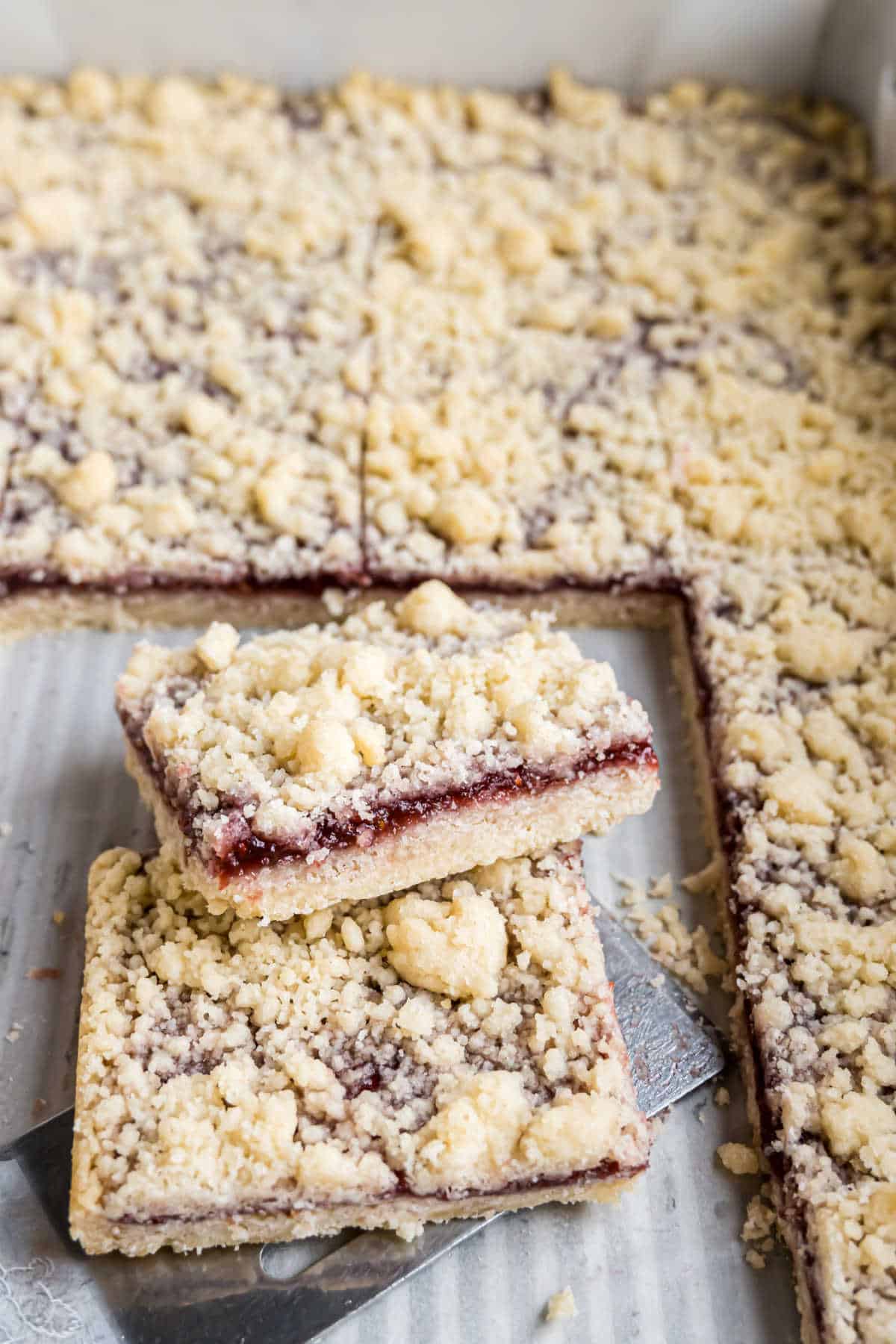 Two raspberry shortbread bars are stacked on a spatula, their crumbly layers tempting, with more bars resting in a baking tray in the background.