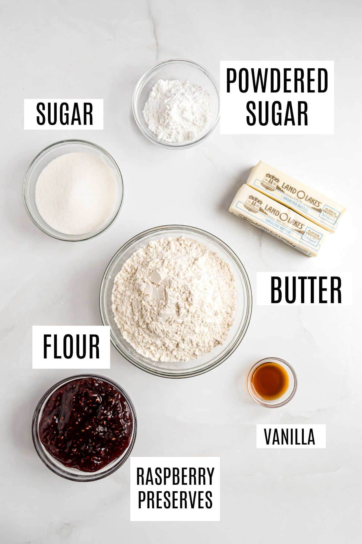 Bowls of sugar, powdered sugar, flour, raspberry preserves, vanilla, and two sticks of butter on a white surface—everything you need to bake delicious raspberry shortbread bars.