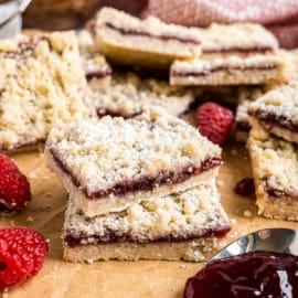 Two stacked raspberry shortbread bars on parchment paper, surrounded by fresh raspberries and a spoonful of jam.