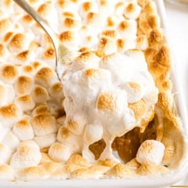 A spoon scoops a serving of sweet potato casserole with marshmallows, lifting the golden, toasted topping from the baking dish.
