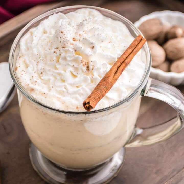 Eggnog with whipped cream and cinnamon stick in glass mug.