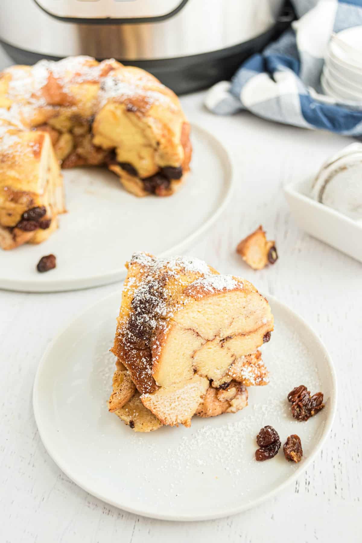 Slice of bread pudding on white plate with pressure cooker in background.