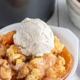 Peach cobbler served in a white bowl with vanilla ice cream on top.