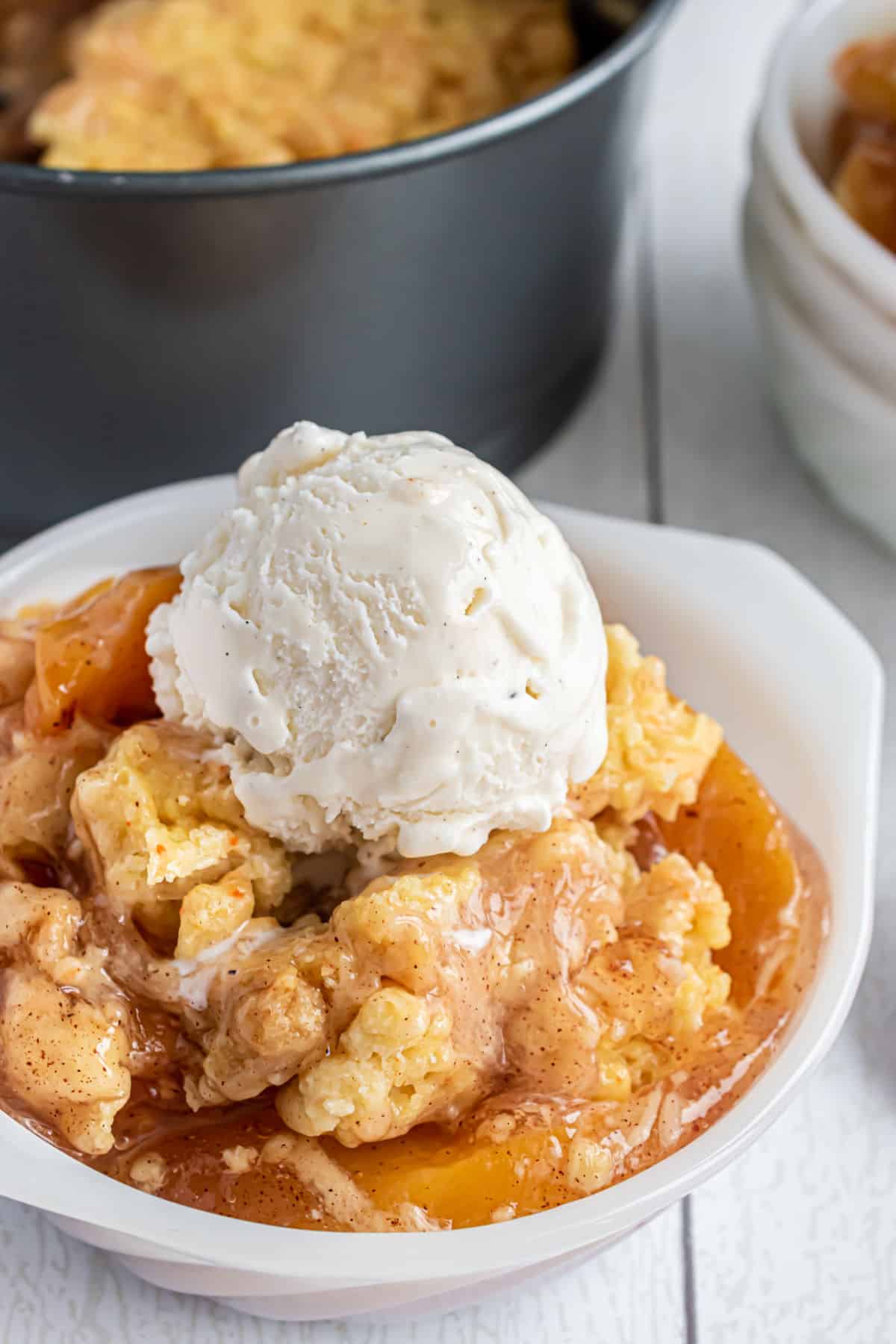 Peach cobbler served in a white bowl with vanilla ice cream on top.