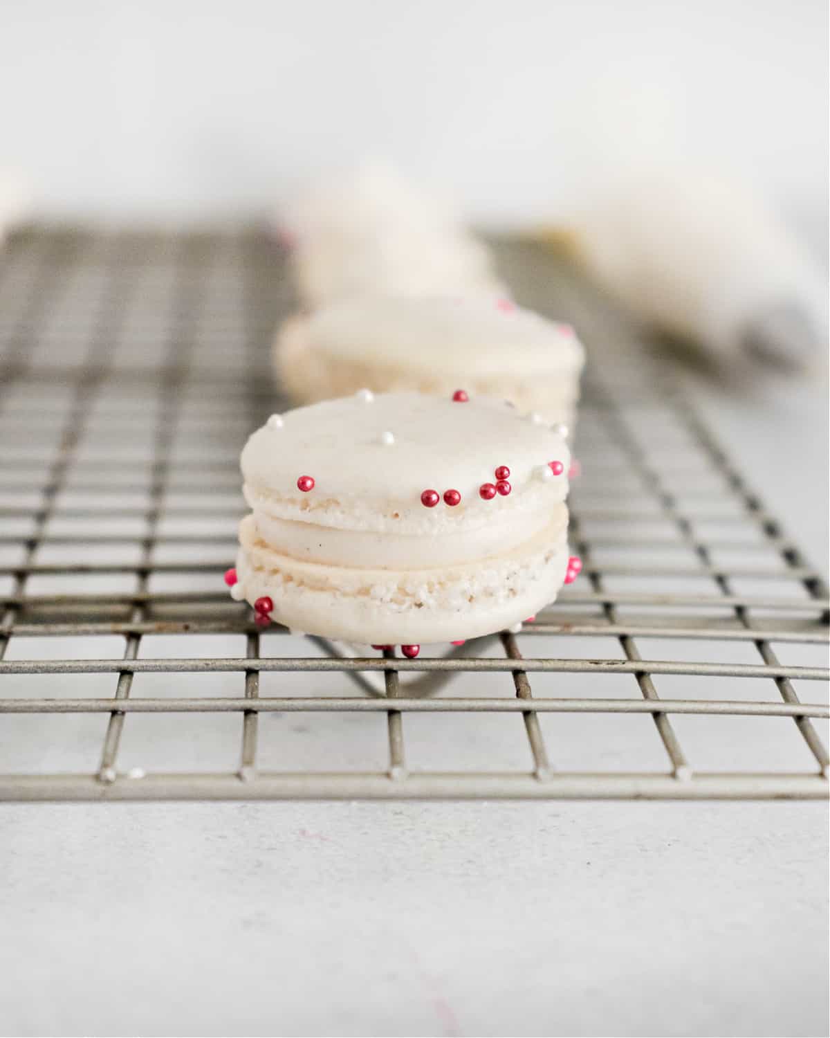 Vanilla bean macarons with pink sprinkles on a wire rack.