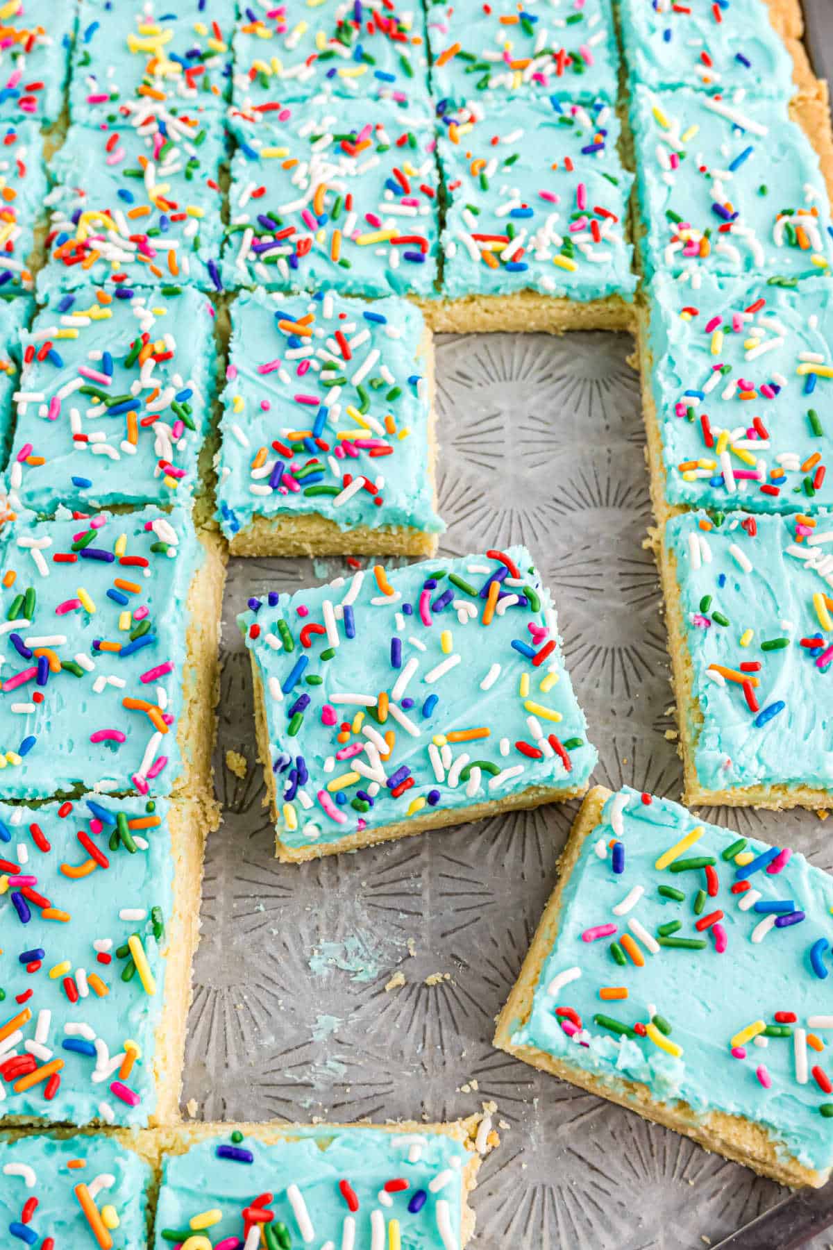 Frosted sugar cookie bars with blue icing and colorful sprinkles are cut into perfect squares on a baking tray, making these frosted sugar cookie bars an irresistible treat for any occasion.