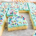 Frosted sugar cookie bars with blue icing and colorful sprinkles are cut into squares on a baking tray.