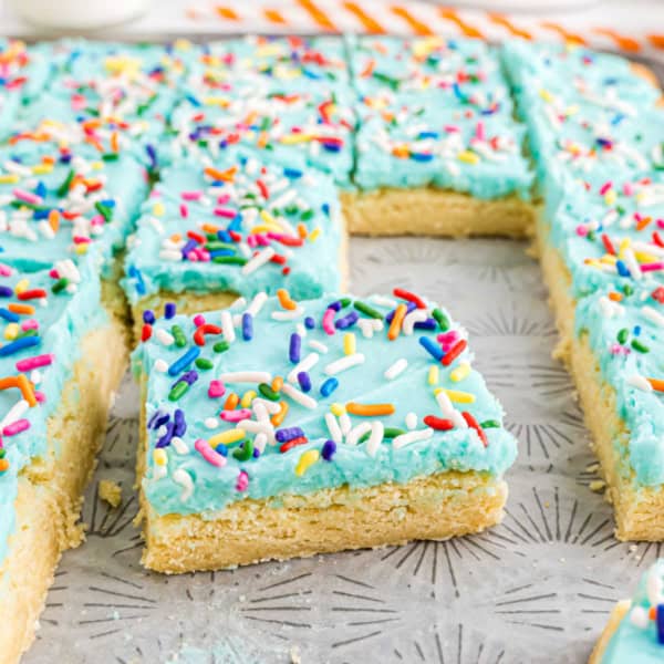 Frosted sugar cookie bars with blue icing and colorful sprinkles are cut into squares on a baking tray.