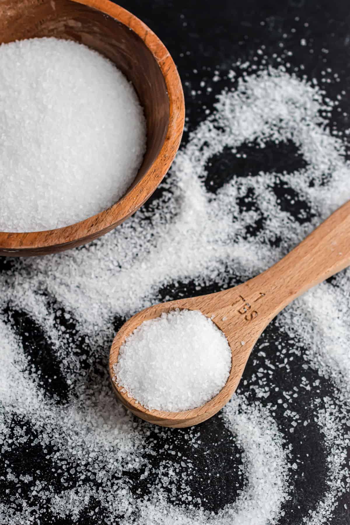 Kosher salt in a wooden bowl spilled onto a balck surface with a wooden spoon.