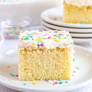 Slice of vanilla cake with vanilla frosting and sprinkles on a white dessert plate.