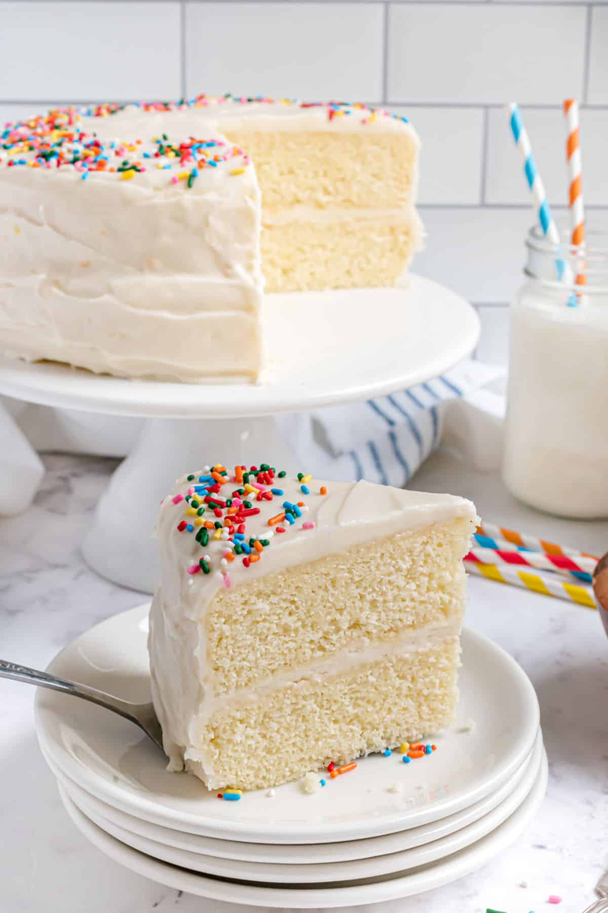 Slice of vanilla layer cake with sour cream frosting and sprinkles on a white plate.