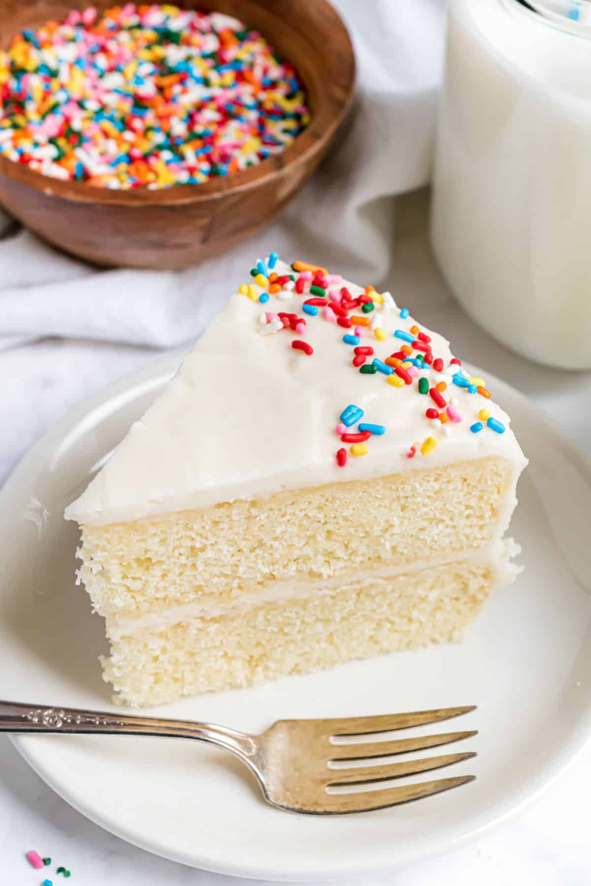 Slice of vanilla cake with sour cream frosting and sprinkles on a white plate with fork.