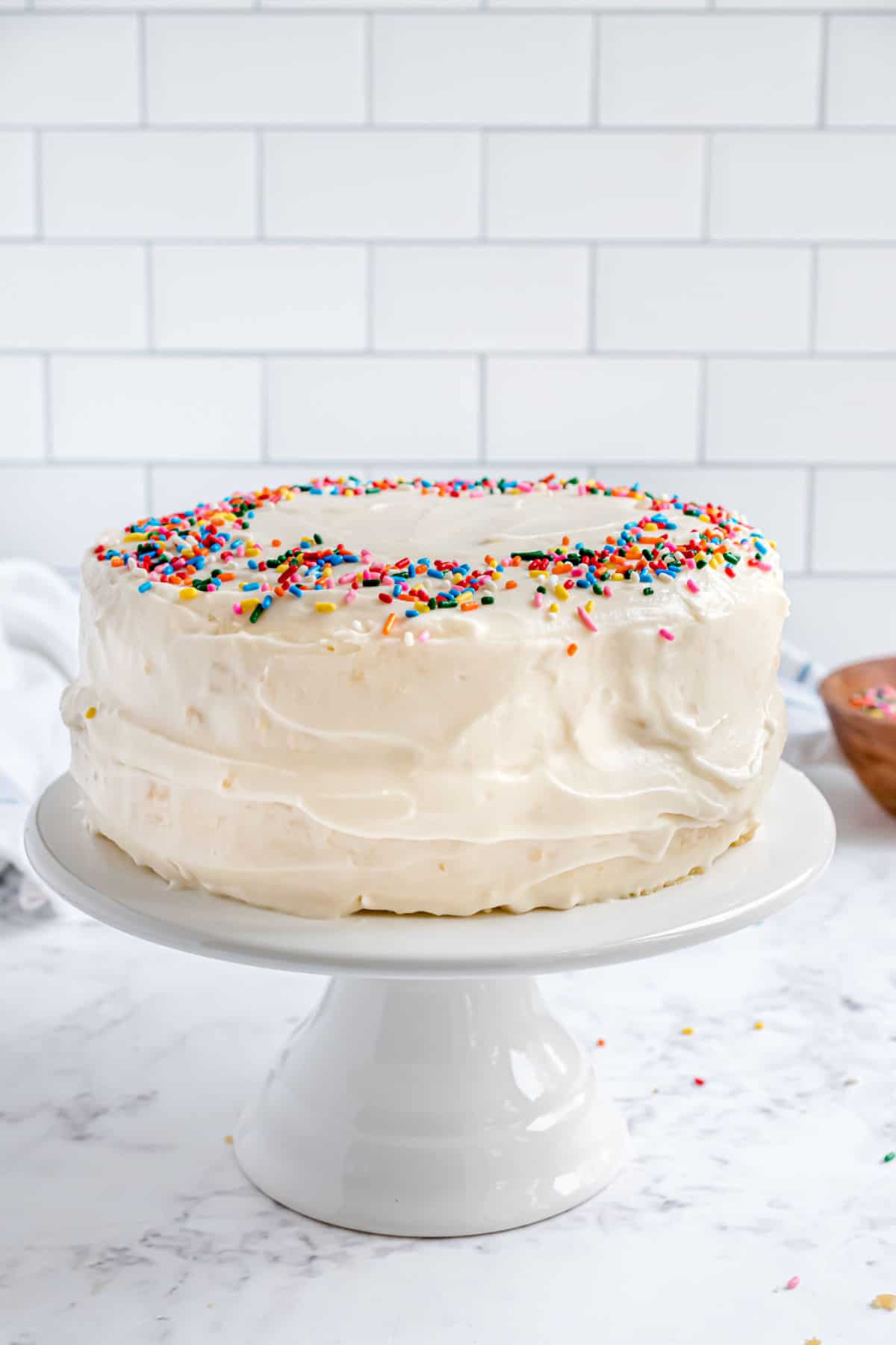 Vanilla cake with vanilla frosting on a white cake stand, topped with sprinkles.