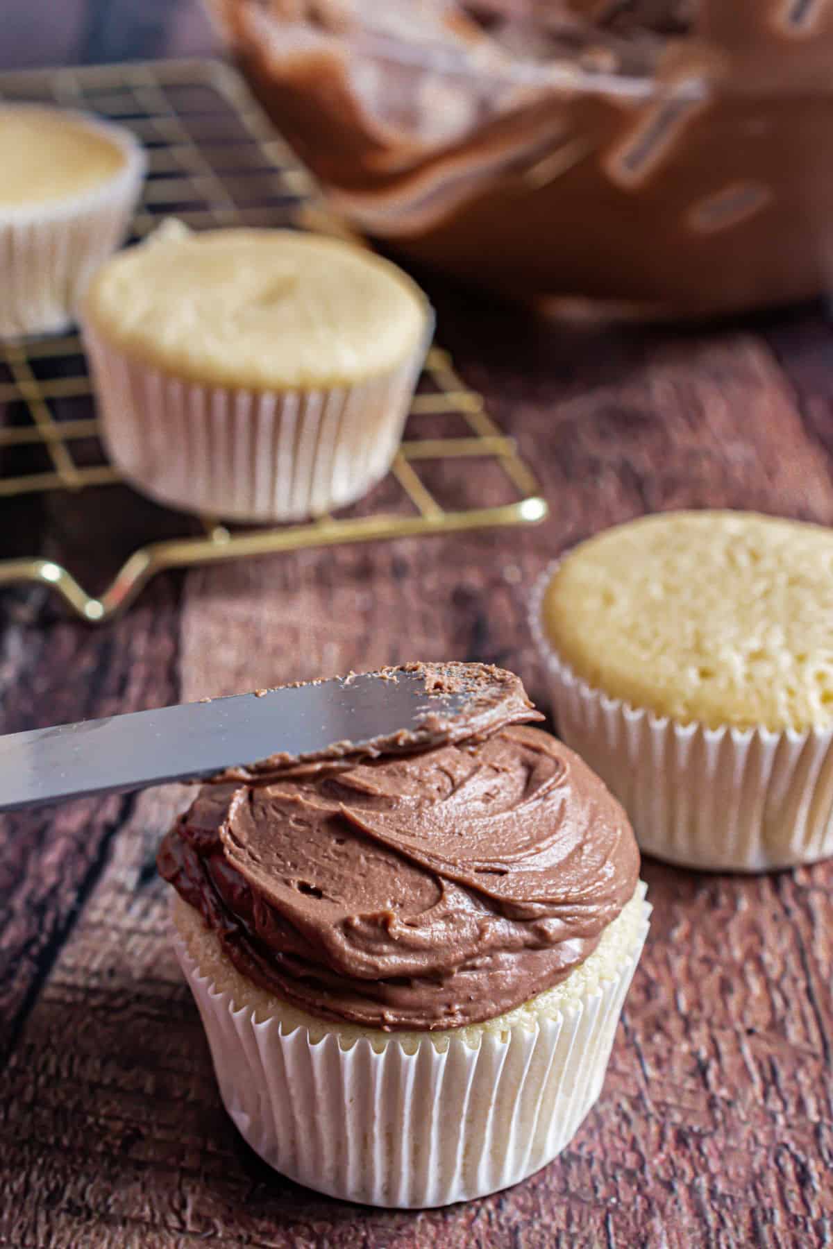 Chocolate sour cream frosting being spread on yellow cupcake with knife. 