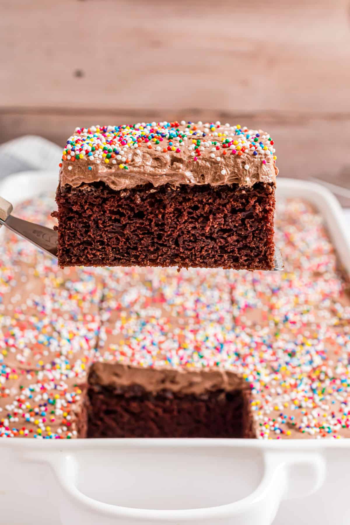 Slice of chocolate cake with frosting being lifted out of a 13x9 dish.