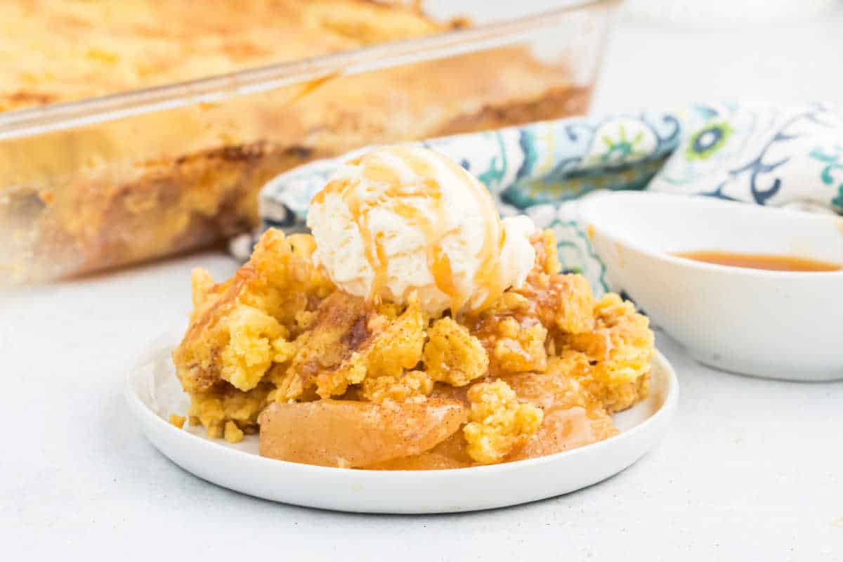A plate of apple cobbler, reminiscent of apple dump cake, topped with vanilla ice cream and caramel sauce, with a baking dish in the background.