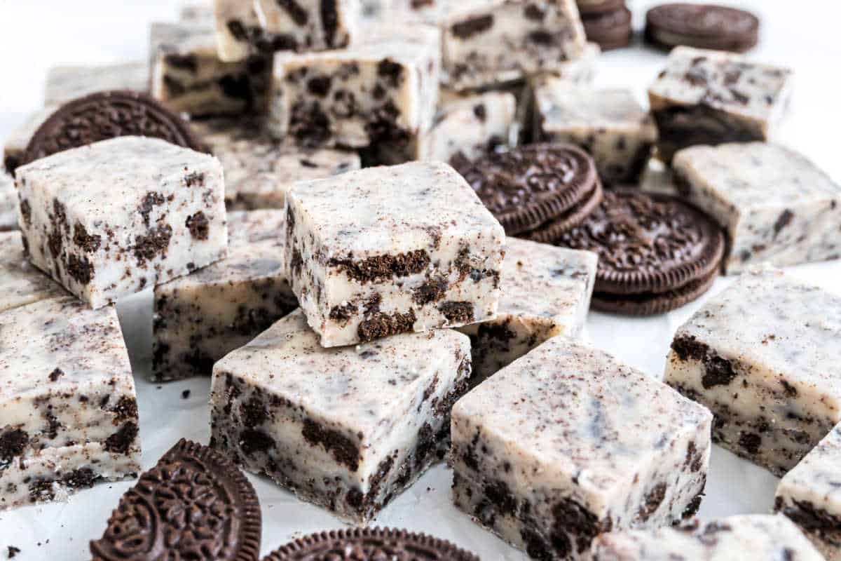 Squares of cookies and cream fudge with Oreo cookie pieces scattered around them on a white surface, highlighting the rich, creamy layers in every bite.
