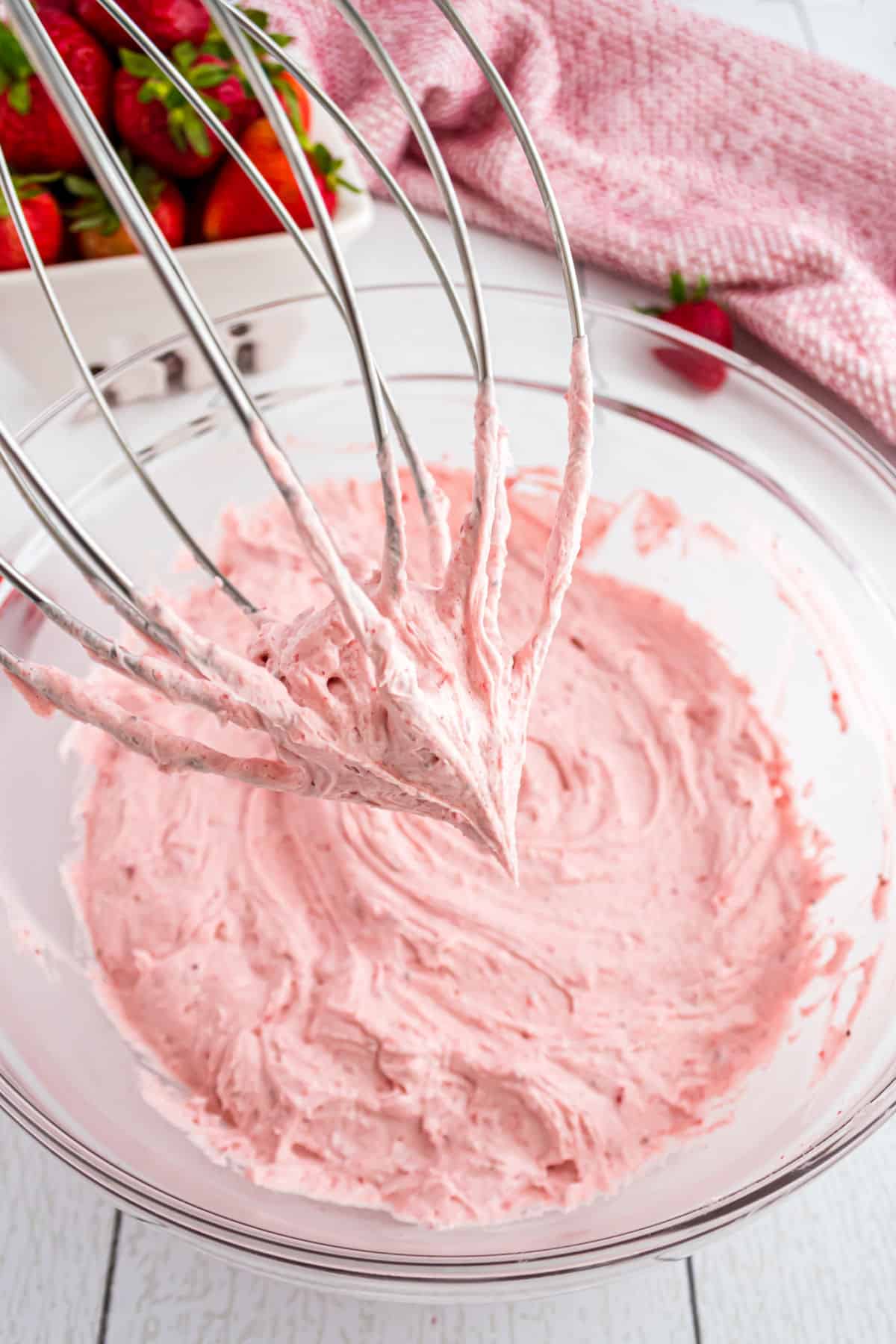 Strawberry frosting in clear glass mixing bowl with whisk attachment.