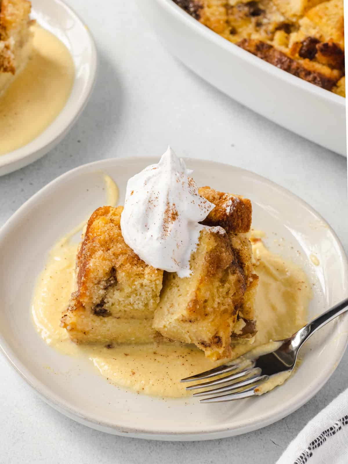 Scoop of apple bread pudding on a plate with vanilla custard and whipped cream.