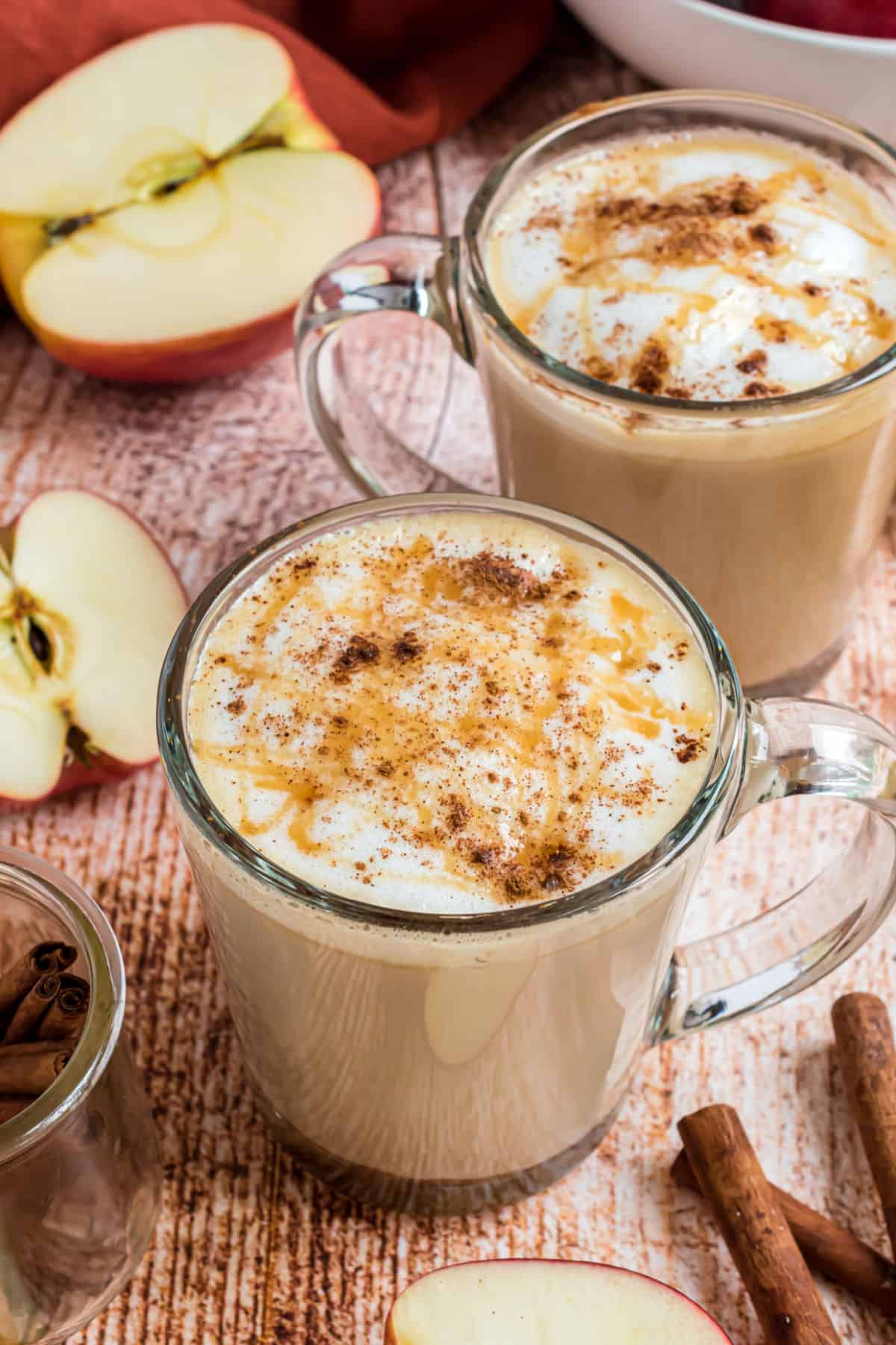 Two clear glass mugs with apple crisp macchiatos topped with caramel syrup.