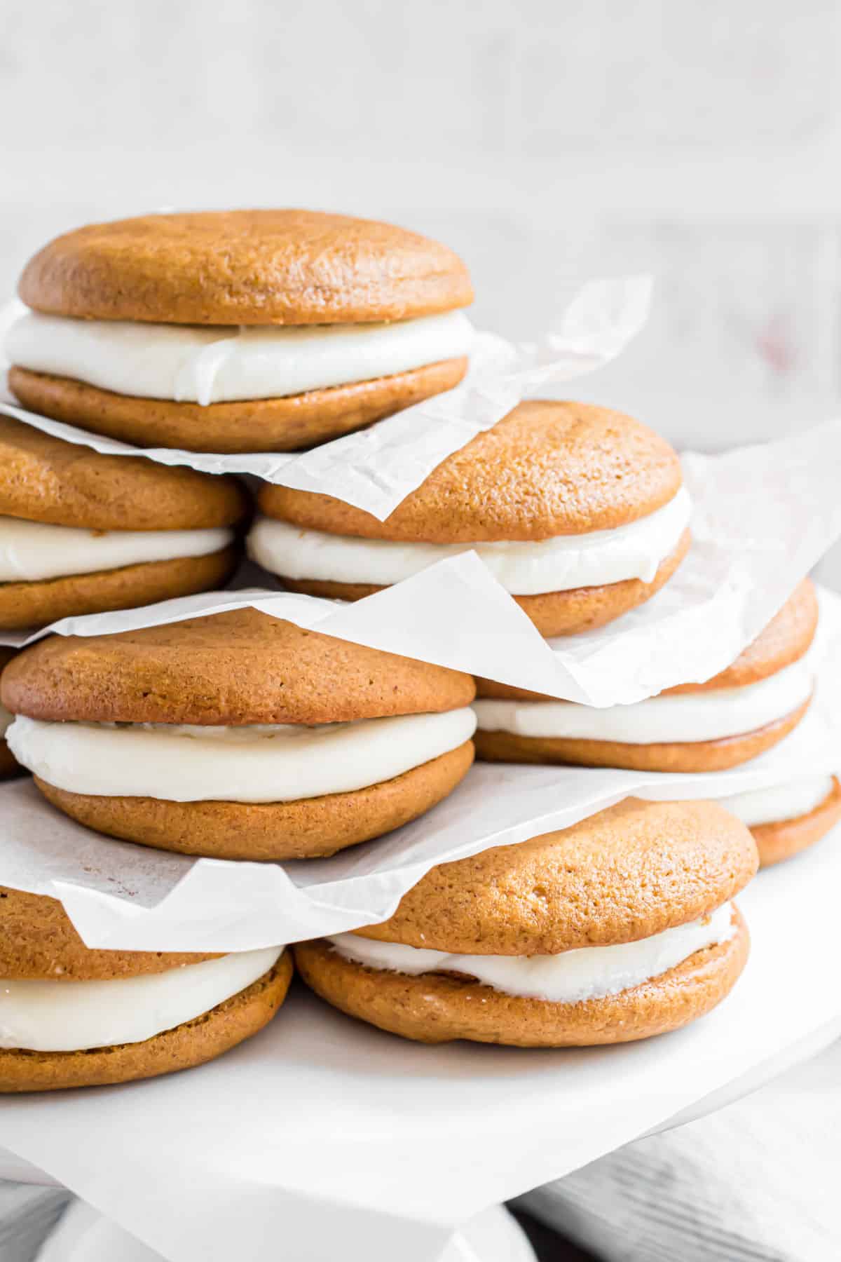 Pumpkin whoopie pies stacked on a cake platter with parchment paper between each cookie.