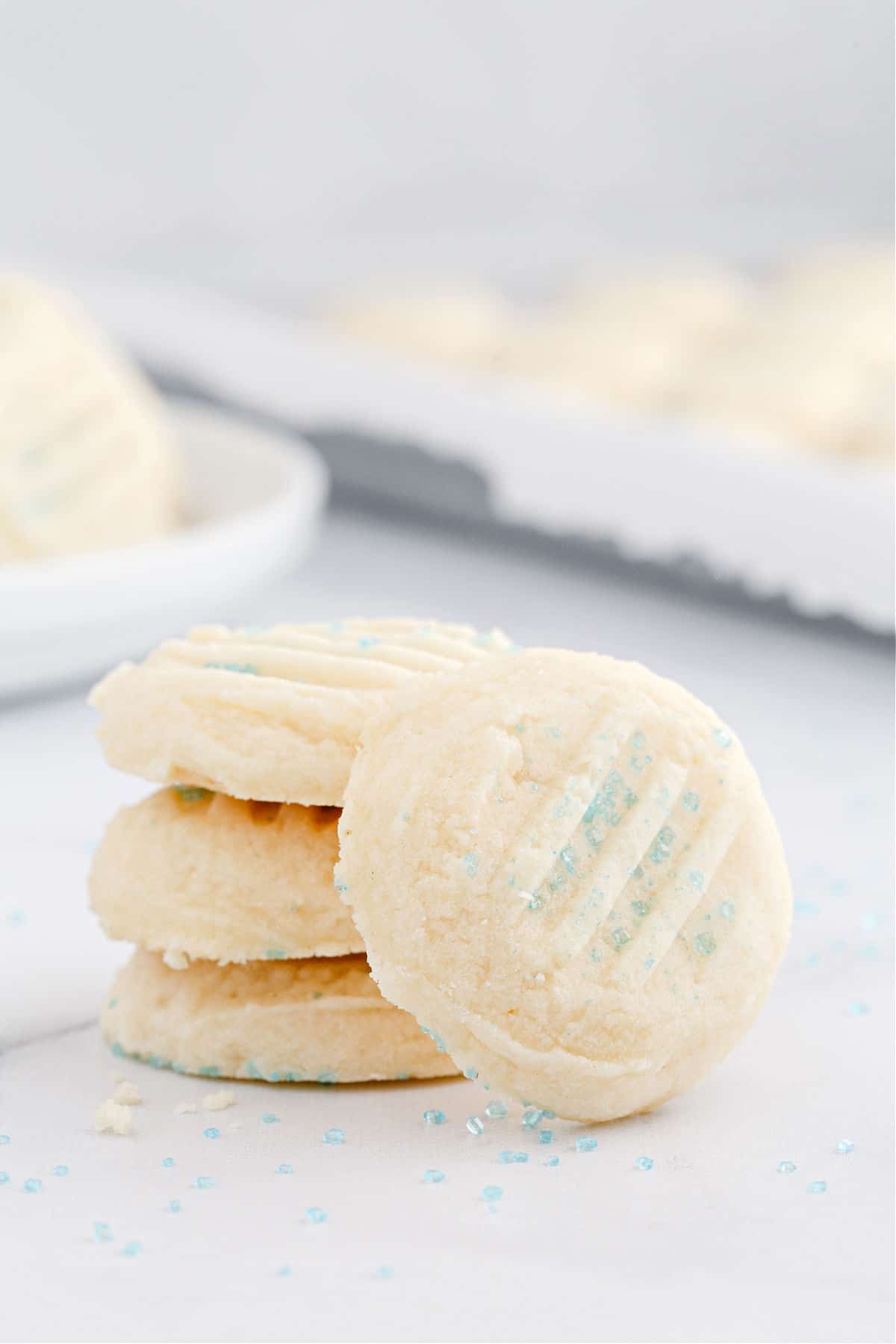 Whipped shortbread cookies stacked on parchment paper with light blue sprinkles.